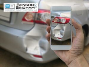 Close-up of a hand holding a smartphone, capturing photographic evidence of a car's rear damage, with Benson Bingham logo visible